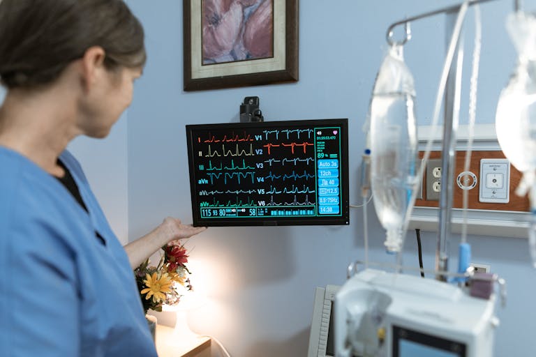 A nurse in blue scrubs examines a medical monitor displaying vital signs in a hospital setting.